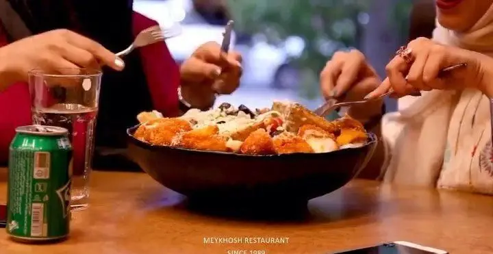 دو مشتری در رستوران میخوش مشغول صرف یک کاسه بزرگ غذای خوشمزه، مانند سالاد ویژه یا سیب زمینی، با نوشیدنی خنک هستند.