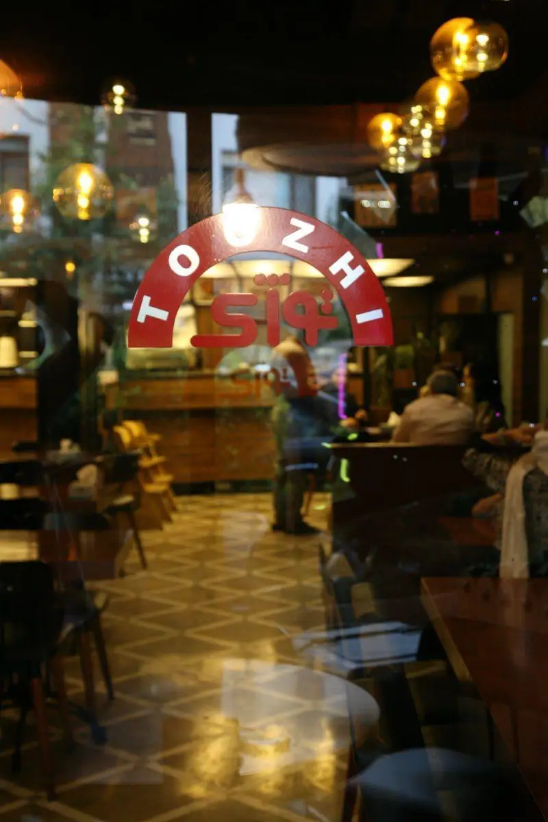 ورودی کافه رستوران توژی با تابلو نام، نمایشگر فضای داخلی دنج، گرم و دلپذیر همراه با میز و صندلی و حضور مشتریان.