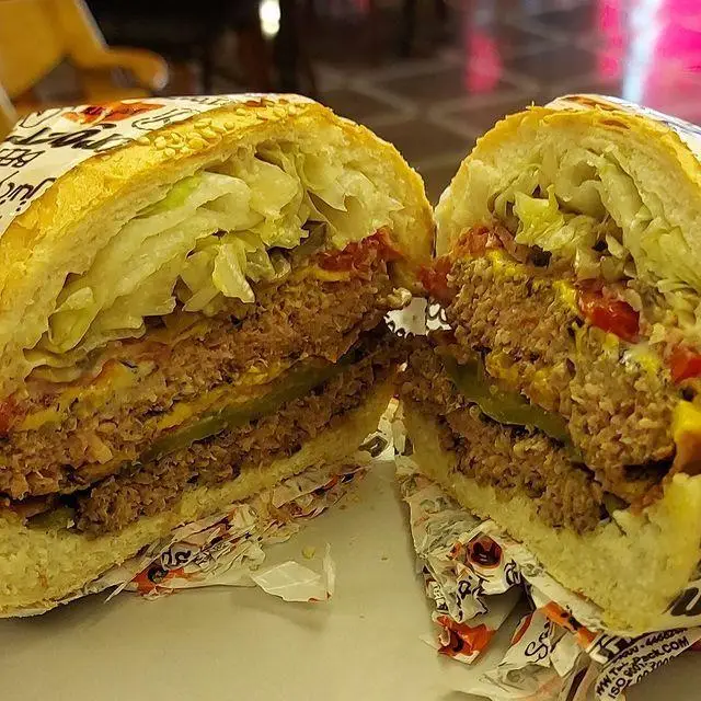 تصویری نزدیک از یک ساندویچ همبرگر گوشت دوبل یا تریپل از کافه رستوران توژی، با نان کنجدی، پنیر، کاهو و خیارشور که به صورت لایه ای چیده شده و آماده سرو است.