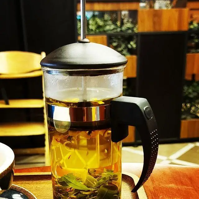 فرنچ پرس شیشه‌ای حاوی چای گیاهی و دمنوش گرم، تجربه‌ای دلپذیر از کیفیت بالا در فضای دنج کافه رستوران توژی.