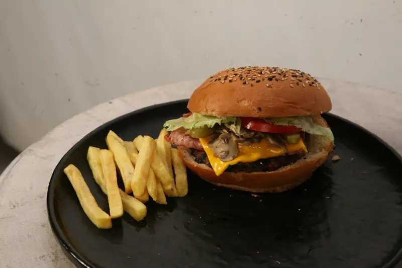 تصویر همبرگر جذاب و پرملات کافه رستوران توژی شامل پنیر، قارچ و بیکن، سرو شده کنار سیب زمینی سرخ کرده خوشمزه.