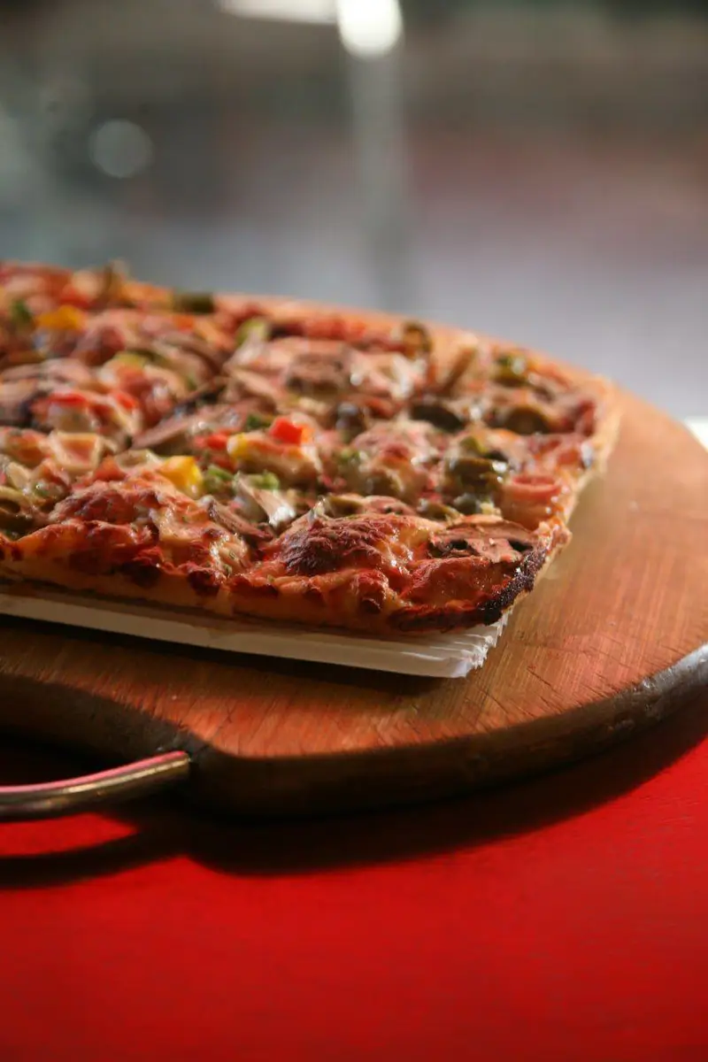 پیتزای تازه و خوشمزه با پنیر طلایی، قارچ و فلفل دلمه‌ای رنگی، آماده سرو روی تخته چوبی در کافه رستوران توژی.