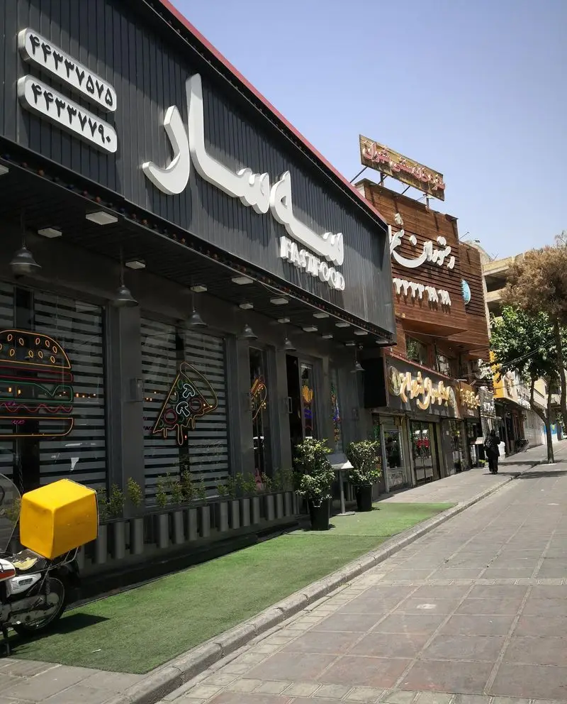 نمای بیرونی فست فود سه سار در تهران با لوگوی نئونی پیتزا و همبرگر، موتورسیکلت دلیوری و شماره‌های تماس ۴۴۳۳۷۵۷۵، ۴۴۳۳۷۷۹۰.
