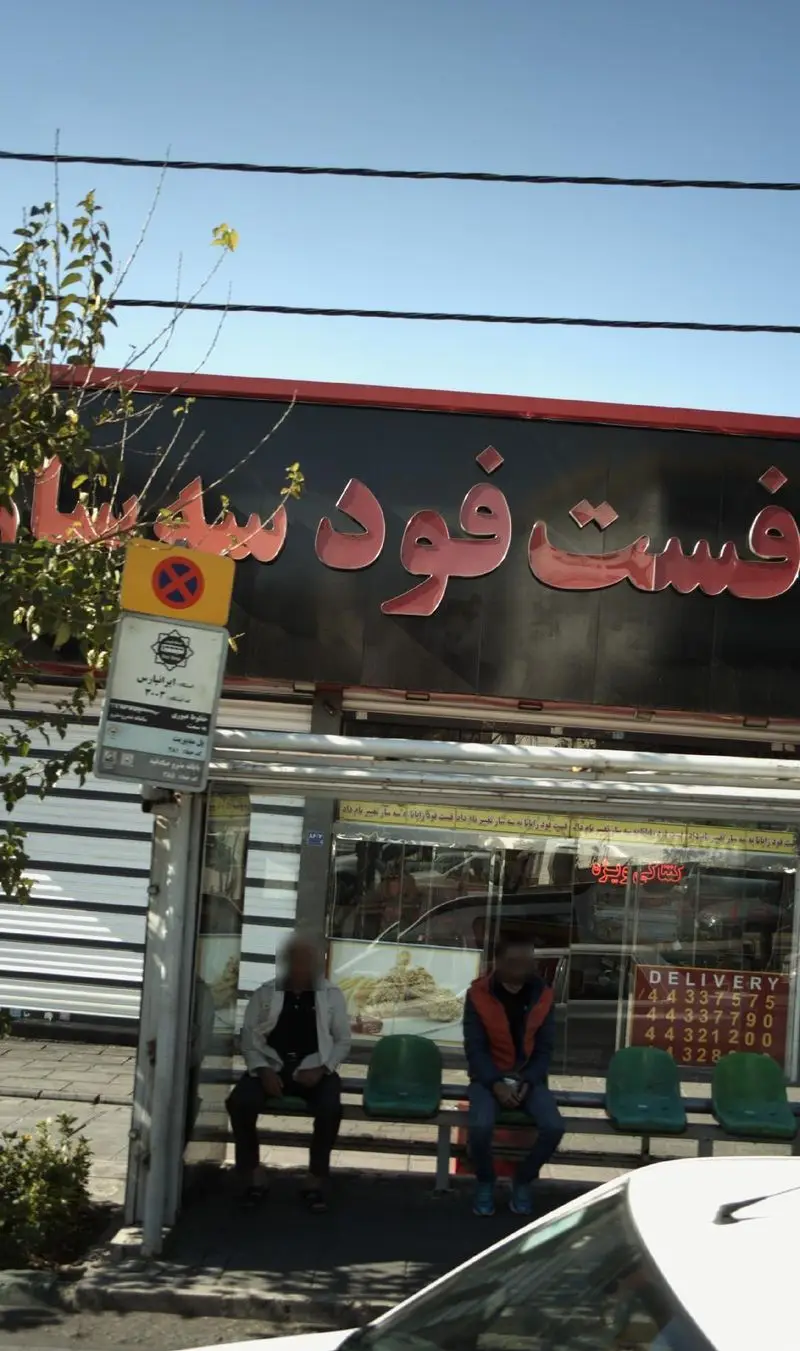 تصویر نمای بیرونی فست فود سه سار، شامل تابلوی درخشان و شماره‌های تماس دلیوری، در کنار یک ایستگاه اتوبوس شلوغ.