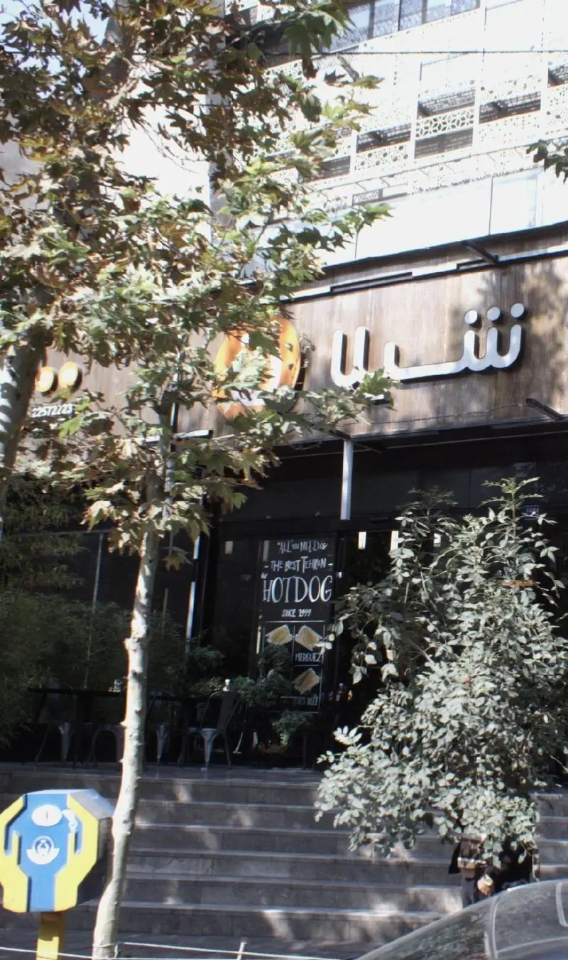 تصویری از نمای بیرونی فست فود شیلا، شعبه دولت تهران، با تابلوی بزرگ شیلا و بنر "بهترین هات‌داگ تهران از سال ۱۹۹۹".