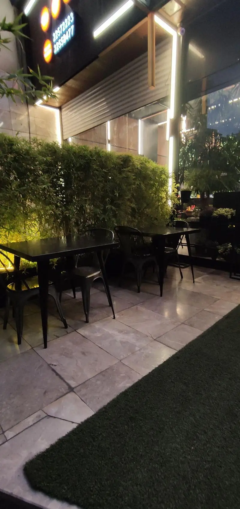 تصویری از نمای شب فضای باز فست فود شیلا شعبه دولت تهران، با میز و صندلی‌های شیک و پرچین سبز زیبا، ایده‌آل برای شام.