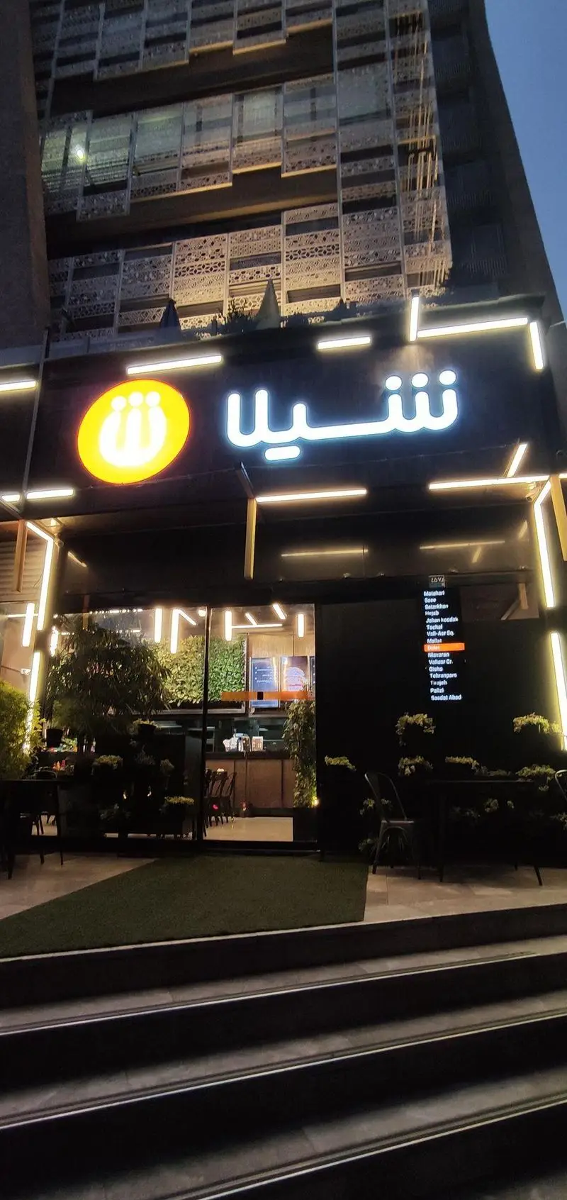 نمایی شبانه از رستوران و فست فود شیلا شعبه دولت با تابلو نئون درخشان، دکوراسیون مدرن، محوطه نشیمن خارجی و فضای سبز زیبا.