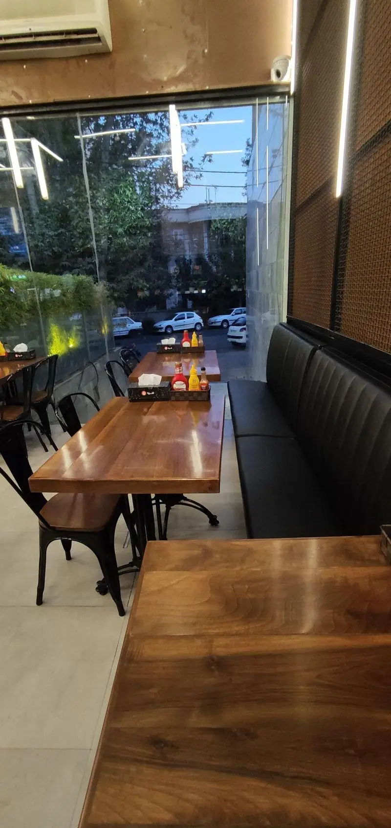 محیط دلنشین فست فود شیلا شعبه دولت با دکوراسیون شیک، میزهای چوبی براق، صندلی‌های مشکی و پنجره‌های وسیع رو به فضای سبز و خیابان.