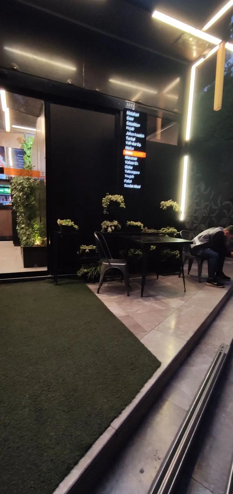 ورودی مدرن فست فود شیلا شعبه دولت در تاریکی شب، با تابلو منو روشن، میز و صندلی‌های فضای باز و دکور گیاهی سبز.