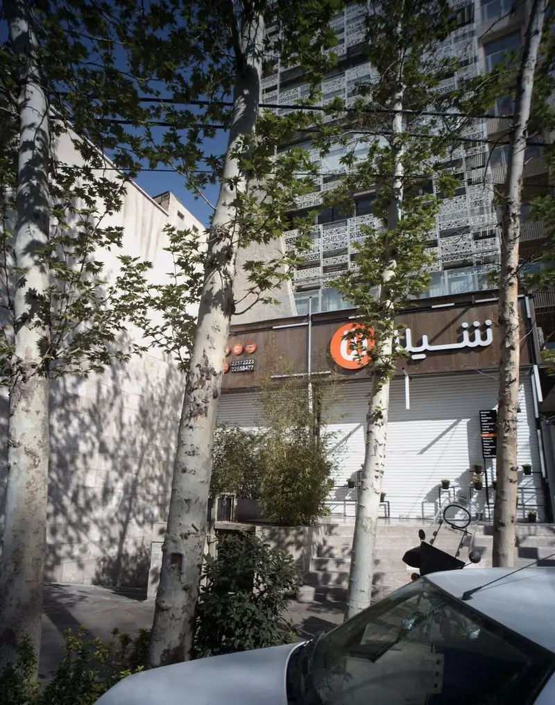 نمای بیرونی فست فود شیلا شعبه دولت در تهران با درختان بلند، نمای ساختمان و تابلوی نارنجی رنگ رستوران در یک روز آفتابی.