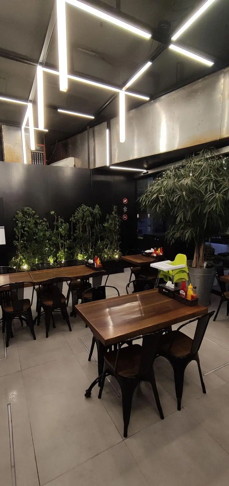 فضای داخلی فست فود شیلا شعبه دولت با نورپردازی خطی مدرن، میز و صندلی‌های چوبی و فلزی، و دکوراسیون سبز و صنعتی.