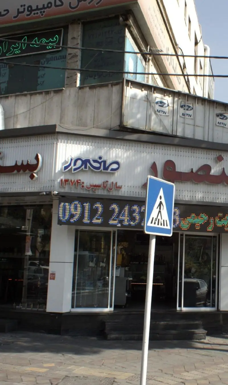 نمای بیرونی مغازه بستنی منصور در روز روشن، با سردر و تابلوی LED که شماره 0912243 و سال تاسیس 1374 را نشان می‌دهد.