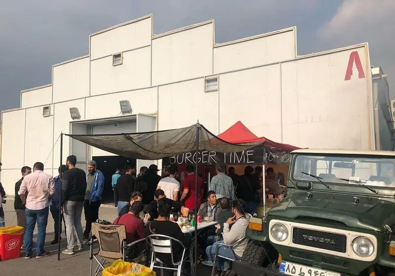 کسب و کار آفرود گریل برگر در فضای باز با مشتریان شاد در حال لذت بردن از غذا، در کنار یک تویوتا لندکروزر سبز رنگ کلاسیک.