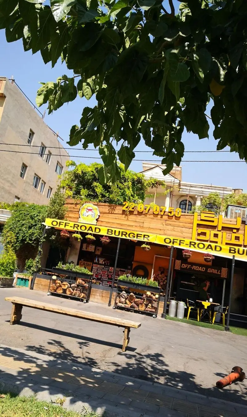 نمای خارجی رستوران آفرود برگر و گریل، با تابلوی زرد، دکوراسیون جذاب چوبی و هیزم، فضای سبز فراوان و نیمکت چوبی.