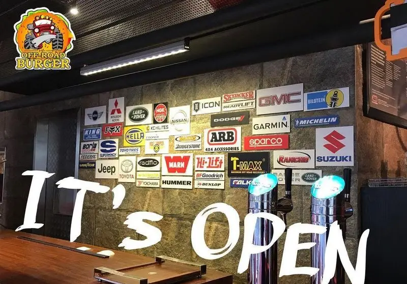 تصویری از دکوراسیون داخلی رستوران آفرود برگر، دیواری با مجموعه‌ای از لوگوهای برندهای خودرویی و آفرود، با پیام "اینجا باز است".