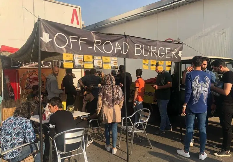 مردم مشغول خرید از فودتراک "آفرود گریل (برگر)" با تابلوی بزرگ "OFF-ROAD BURGER" و منوهای آویزان زرد در محیطی شلوغ.