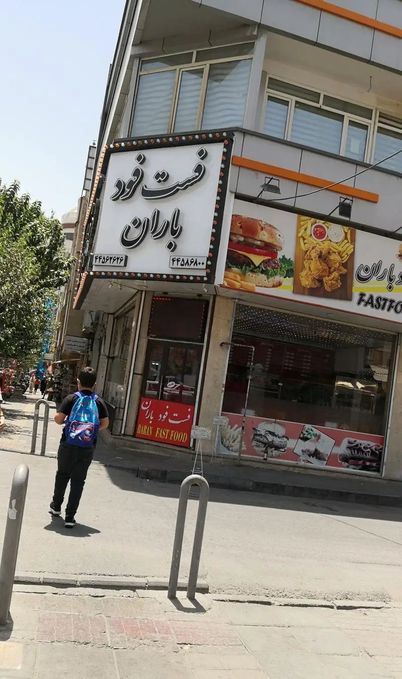 تصویری از نمای بیرونی فست فود باران با تابلوی بزرگ حاوی نام و شماره تماس، نمایش منوی جذاب شامل همبرگر و مرغ سوخاری به همراه سیب زمینی.