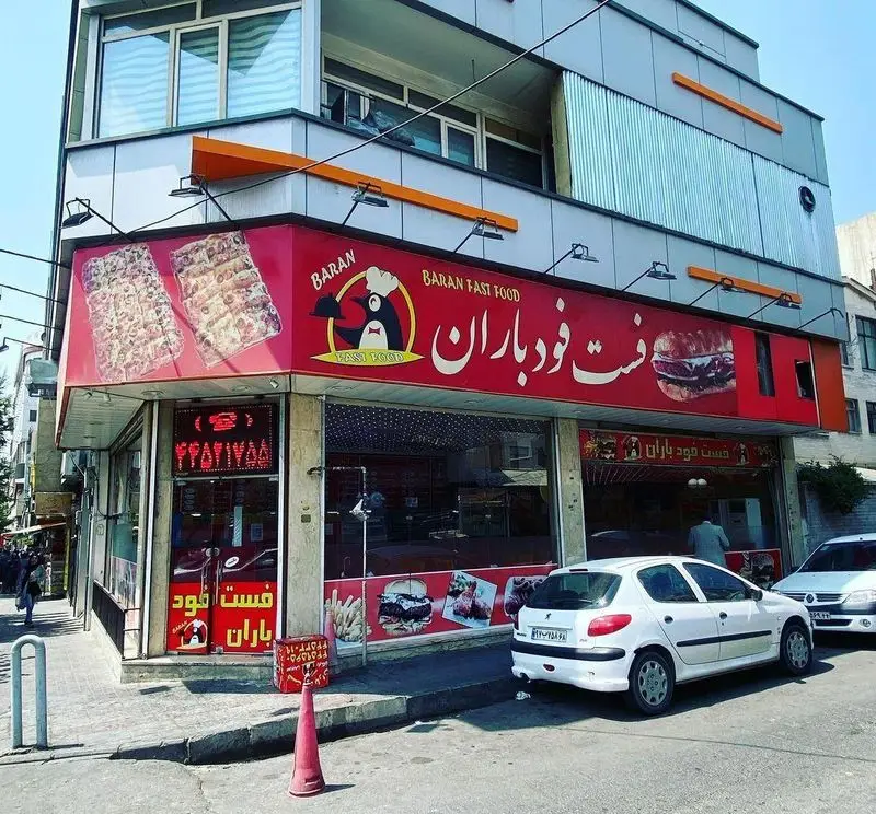 نمای بیرونی فست فود باران، تابلوی قرمز بزرگ با لوگوی پنگوئن و تبلیغ پیتزا و همبرگر. خیابان شلوغ در شهر تهران.