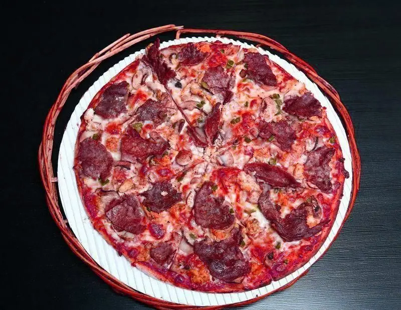 پیتزای بزرگ و خوش‌رنگ رستوران ایتالیایی ژولی با گوشت فراوان، قارچ و فلفل، روی سینی سفید با حاشیه حصیری.