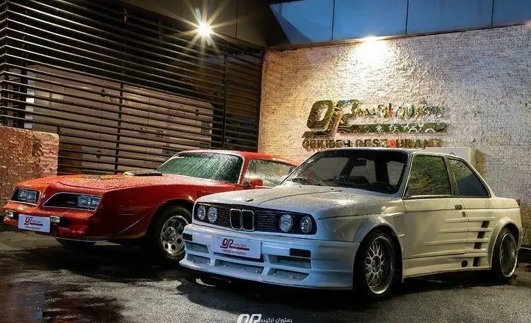 تصویر شبانه چلوکبابی ارکیده با زمین خیس، نمایش خودروهای کلاسیک پونتیاک فایربرد قرمز و بی ام و E30 سفید روبروی ورودی.