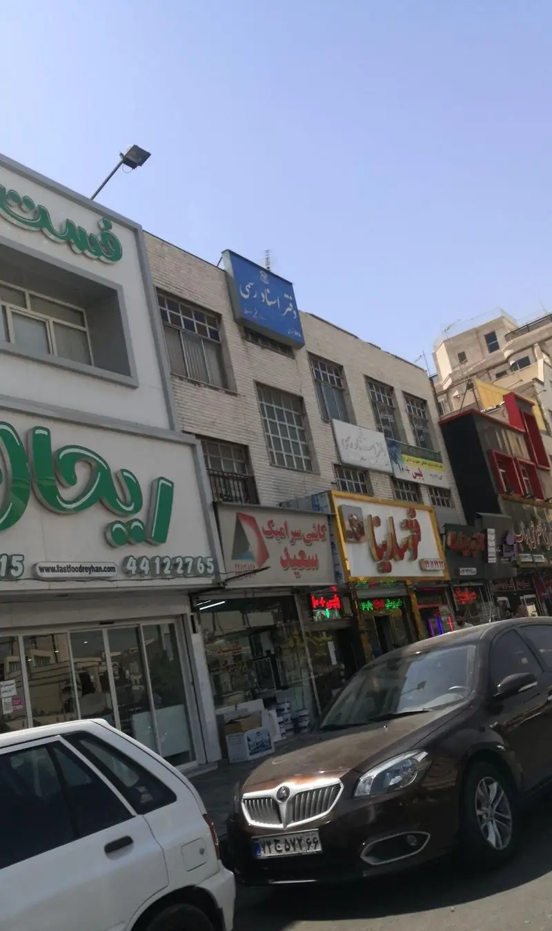 نمایی از فست فود ریحان در یک روز آفتابی در خیابان شلوغ شهری با تابلو سبز، آدرس وبسایت www.fastfoodreyhan.com و تلفن ۴۴۱۲۲۷۶۵.