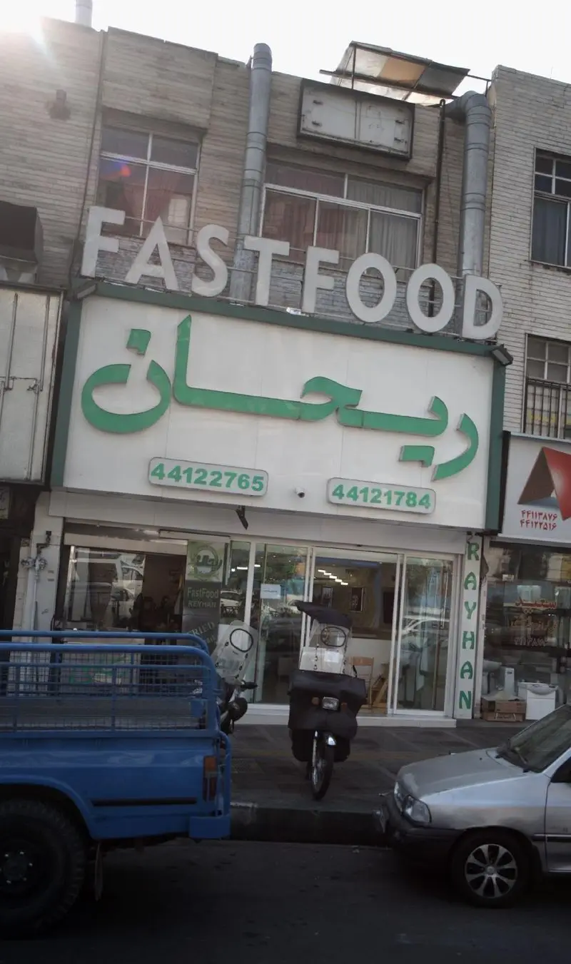 نمای بیرونی فست فود ریحان با تابلو Fast Food و شماره تلفن ۴۴۱۲۲۷۶۵، ۴۴۱۲۱۷۸۴ در خیابان. یک وانت و موتور در جلوی مغازه پارک شده‌اند.