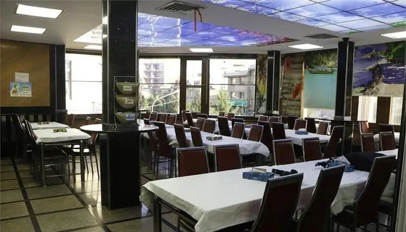 سالن غذاخوری بزرگ فست فود اغذیه نشاط با میزهای سفید و صندلی‌های راحت، آماده پذیرایی از مشتریان با چشم‌انداز شهری دلنشین و دکوراسیون زیبا.