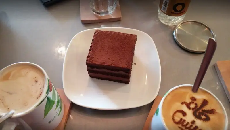 کافه رئیس: یک میز پر از لذت شامل دو فنجان قهوه لاته گرم، یکی با طرح کافه رئیس و دیگری با طرح برگ، به همراه کیک شکلاتی غلیظ و دلپذیر.