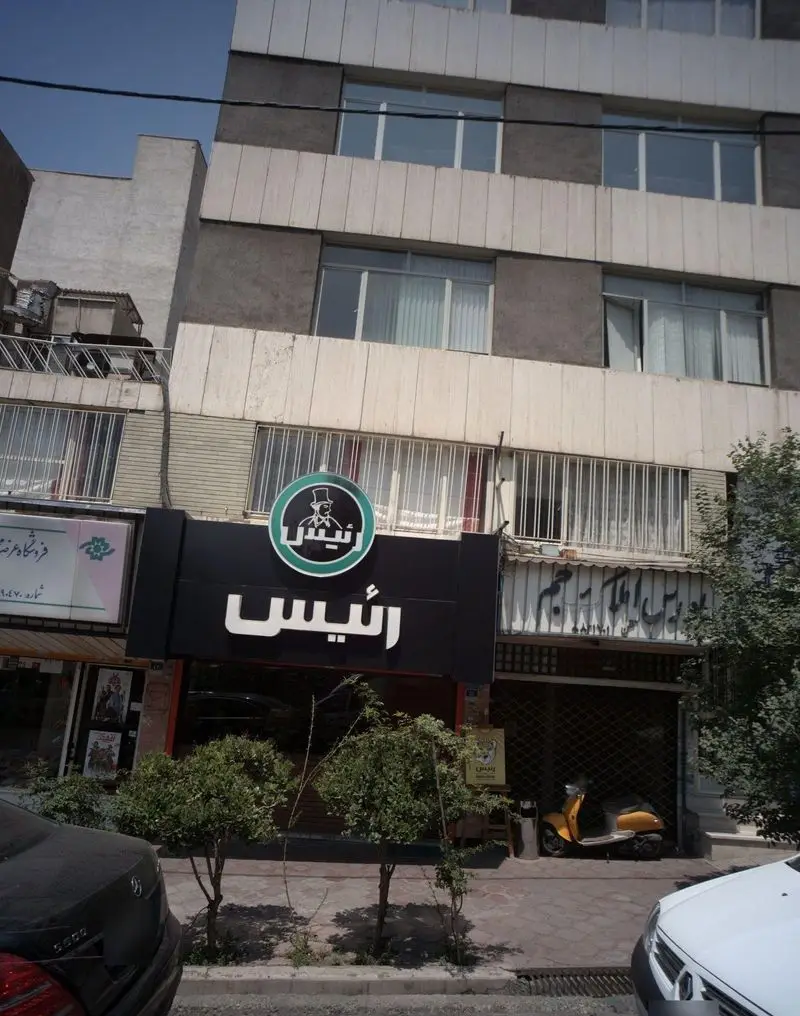 عکس نمای بیرونی کافه رئیس، تابلوی مشکی با لوگوی مرد کلاهدار، شامل ساختمان، خیابان، ماشین‌ها. محیطی دلنشین برای قهوه و دورهمی.