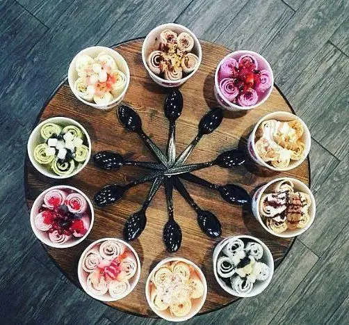 کافه بستنی نیترولی: چیدمان زیبا و جذاب از ۱۲ کاسه بستنی رول شده با طعم های متنوع میوه ای و شکلاتی روی میز چوبی.