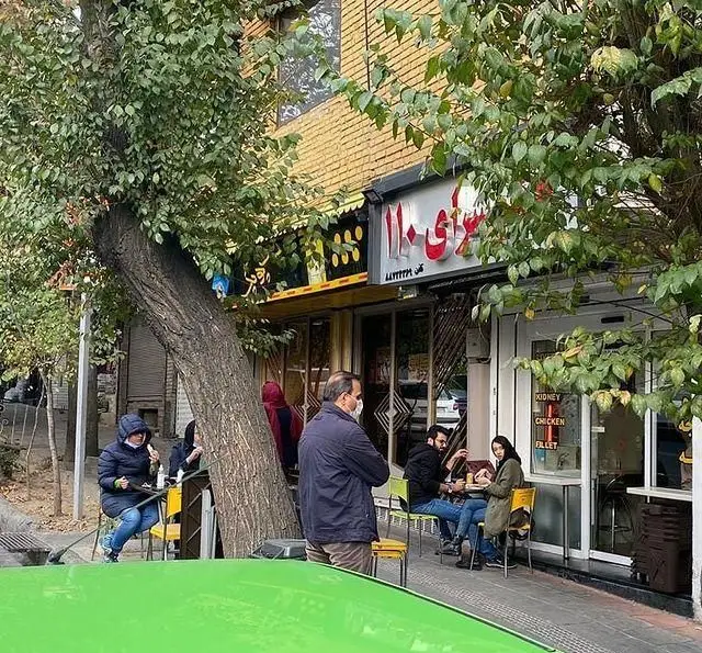 تصویری از نمای بیرونی جگر سرای ۱۱۰، رستورانی معروف در تهران. مشتریان در فضای باز و کنار درخت مشغول خوردن جگر و انواع کباب هستند.