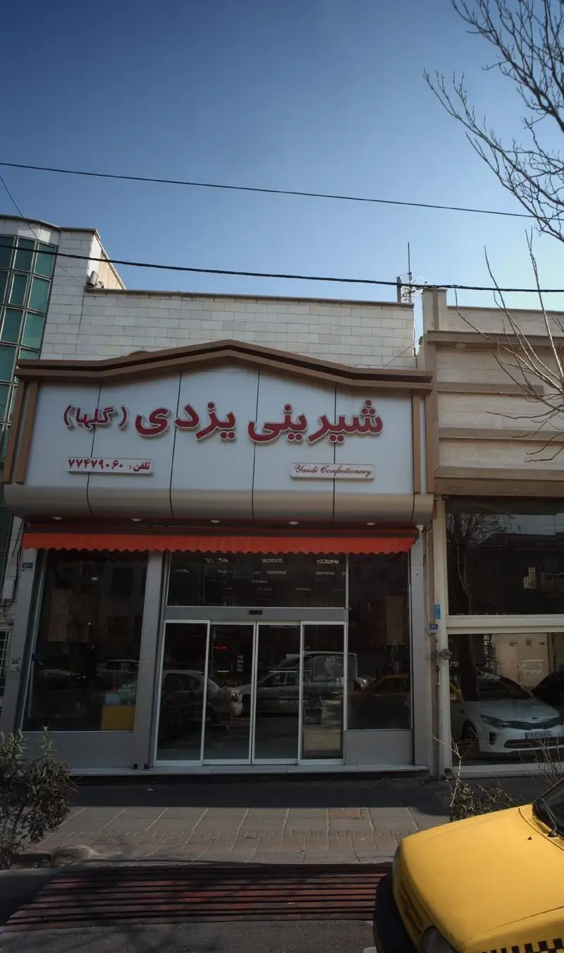 نمای بیرونی قنادی شیرینی یزدی (گلها) با تابلوی بزرگ و روشن، درب‌های شیشه‌ای، مکانی ایده‌آل برای خرید شیرینی سنتی یزدی.