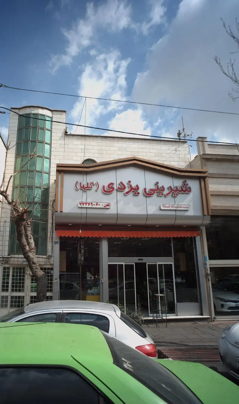 نمای بیرونی قنادی شیرینی یزدی گلها با تابلوی روشن و نام فارسی و انگلیسی، شماره تماس ۷۷۲۷۹۰۶۰، ویترین شیشه‌ای و خودروهای پارک شده در جلو.
