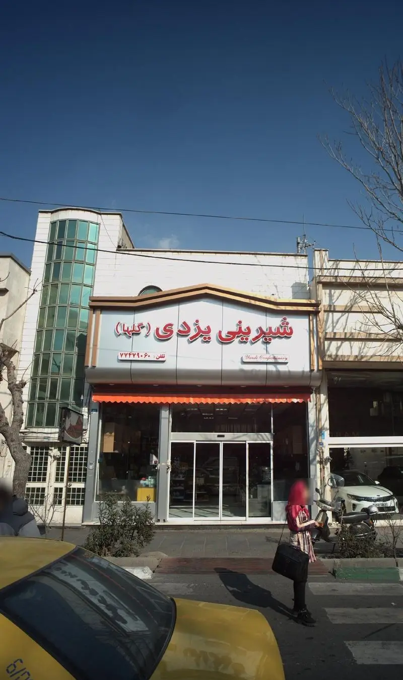 نمای جلوی مغازه شیرینی یزدی گلها با تابلوی درخشان و نام "Yazdi Confectionery"، تلفن ۷۷۴۷۹۰۶۰، زیر آسمان آبی تهران در روز آفتابی.