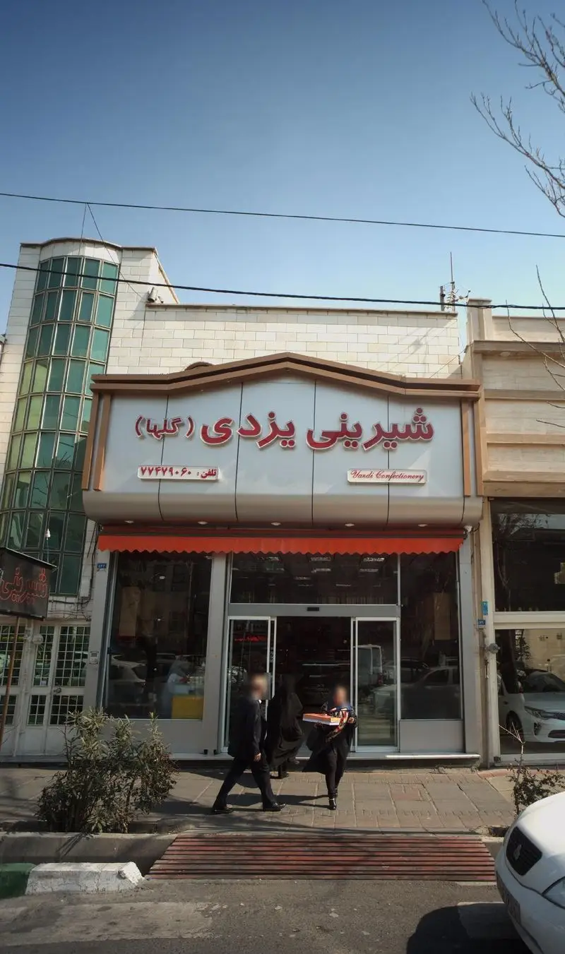 نمای بیرونی قنادی شیرینی یزدی گلها با تابلو قرمز و نمای شیک. مشتریان در حال ورود به فروشگاه برای خرید شیرینی‌های سنتی و خوشمزه یزدی.
