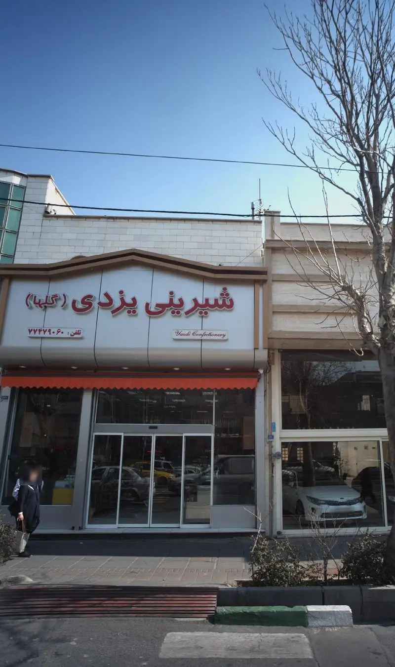 فروشگاه شیرینی یزدی (گلها) با نمای شیک و مدرن، تابلوی قرمز رنگ و درب ورودی شیشه ای در یک خیابان شلوغ شهری.
