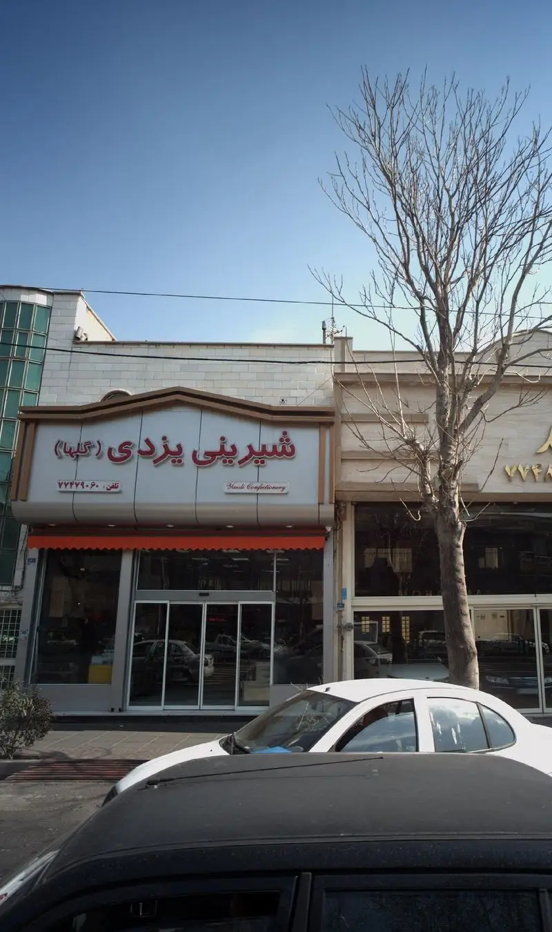 تصویر تابلوی شیرینی یزدی گلها در تهران، قنادی معروف با نمای شیک، ورودی اتوماتیک و چتر آفتابگیر نارنجی.