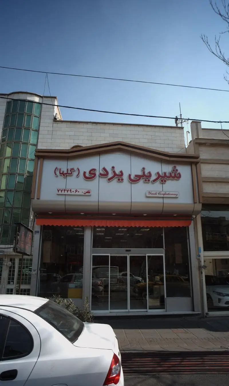 نمای بیرونی قنادی شیرینی یزدی (گلها) با تابلوی روشن حاوی نام فارسی و انگلیسی، درب‌های شیشه‌ای ورودی و خودروی سفید مقابل فروشگاه.