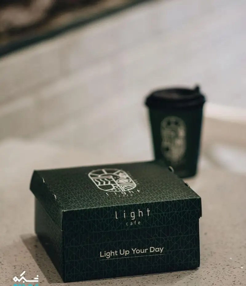 بسته بندی شیک کافه لایت شامل جعبه‌ای سبز تیره با لوگو، شعار "Light Up Your Day" و لیوان قهوه هماهنگ.