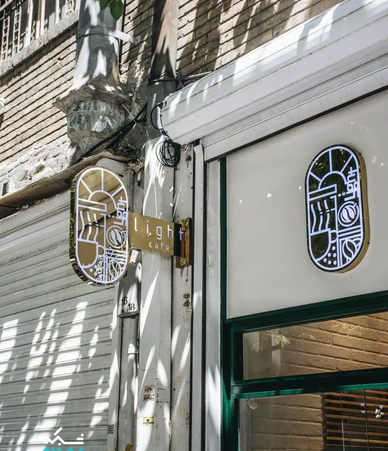 تصویری از ورودی کافه لایت با دو تابلو نئونی مدرن و آرم خاص، نمای خیابانی با سایه روشن آفتاب و معماری شهری.