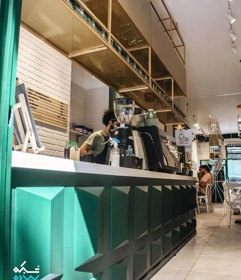 فضای داخلی مدرن کافه لایت، با میز بار سبز رنگ و دکوراسیون شیک طلایی. باریستای حرفه‌ای در حال آماده‌سازی قهوه و نوشیدنی‌های خوشمزه. محیطی دنج برای یک کافی شاپ عالی.