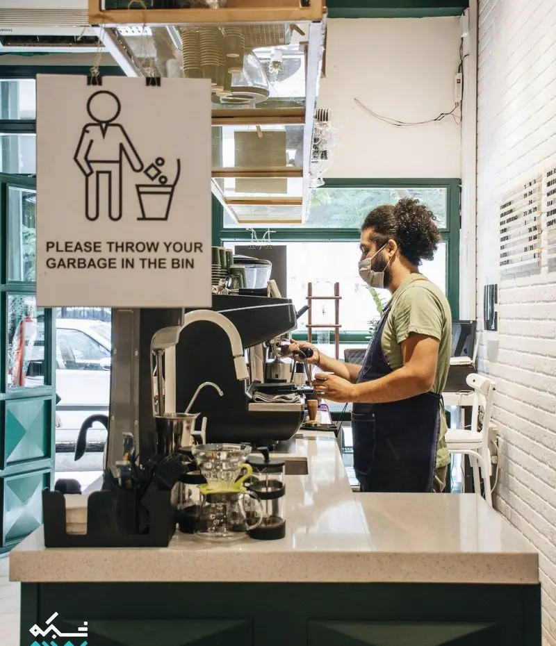 باریستای کافه لایت با ماسک و پیشبند در حال تهیه قهوه با دستگاه اسپرسو. محیط داخلی روشن و تمیز کافه با دکوراسیون مدرن و رعایت بهداشت.