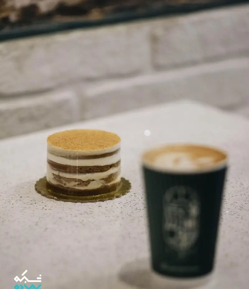 کیک لایه ای کرم‌دار با پودر بیسکویت به همراه قهوه لاته در کاپ مخصوص کافه لایت، انتخابی عالی برای یک استراحت دلپذیر.