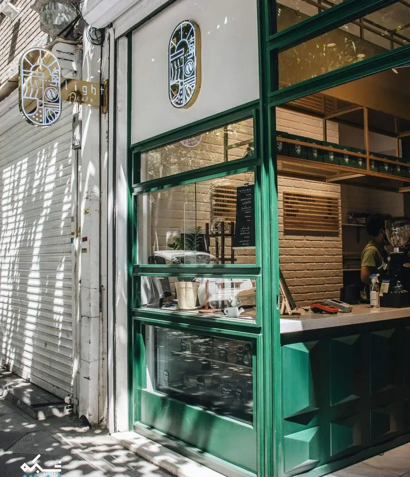 کافه لایت: نمای خارجی شیک با طراحی سبز رنگ، ویترین‌های بزرگ شیشه‌ای و تابلوهای هنری مدرن. نشان Light cafe به وضوح دیده می‌شود.