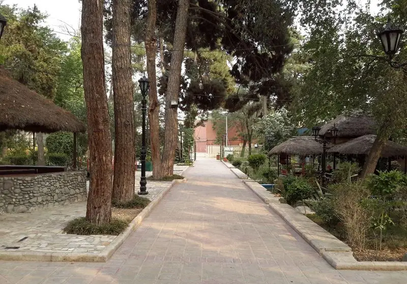 فضای سرسبز و دلنشین رستوران اشراق با مسیر پیاده‌روی سنگفرش، درختان بلند و آلاچیق‌های سنتی برای تجربه‌ای آرامش‌بخش.