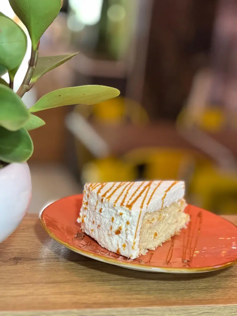 برش کیک خوشمزه کافه پارس با خامه سفید و تزیین کارامل روی بشقاب نارنجی در محیطی دلنشین. انتخابی عالی برای دسر در کافه.