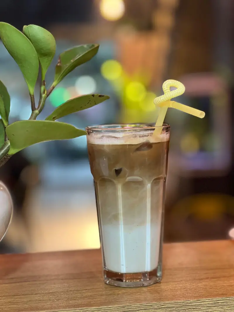 یک لیوان آیس لاته خوش‌رنگ با لایه‌های قهوه و شیر، همراه با نی زرد، در محیط آرام و دلپذیر کافه پارس برای تجربه لحظاتی خوش.