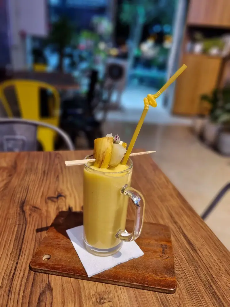 تصویری از یک اسموتی میوه ای خنک و خوشرنگ (موز) در لیوان شیشه ای روی میز چوبی کافه پارس، آماده برای سرو در محیط دلنشین.