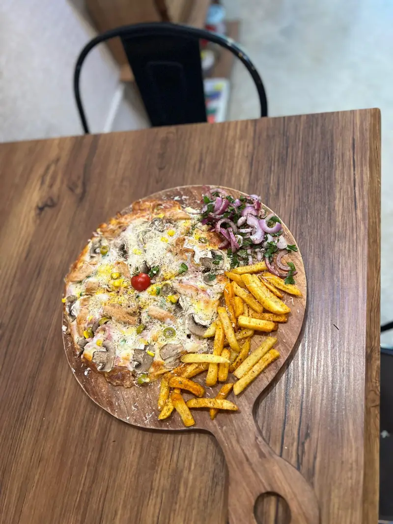 پیتزای خوش طعم و وسوسه‌انگیز کافه پارس شامل قارچ، مرغ و ذرت، همراه با سیب‌زمینی سرخ کرده و سالاد پیاز روی تخته سرو چوبی.