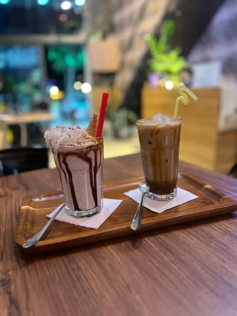 نوشیدنی‌های محبوب کافه پارس: یک میلک شیک شکلاتی با تزئینات و یک قهوه سرد یخی روی سینی چوبی در فضای آرام کافه.